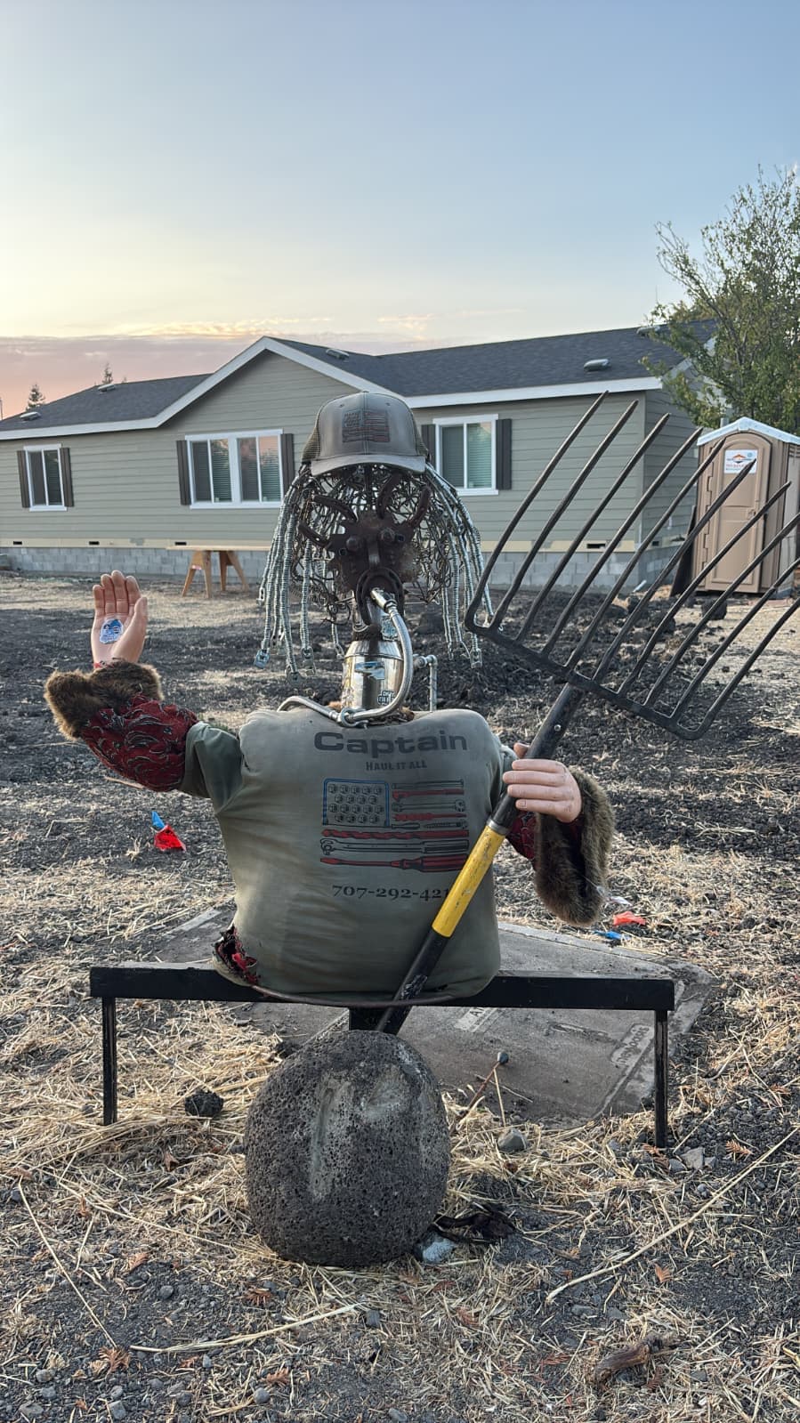 Back view of handmade scarecrow decoration installed in dirt lot near residential home.